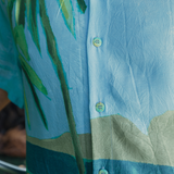 Close-up of the shirt's fabric and buttons. The light blue fabric shows part of the mountain and palm tree print. The buttons are small, round, and blue green with a subtle speckled pattern, blending with the shirt's tropical tones.