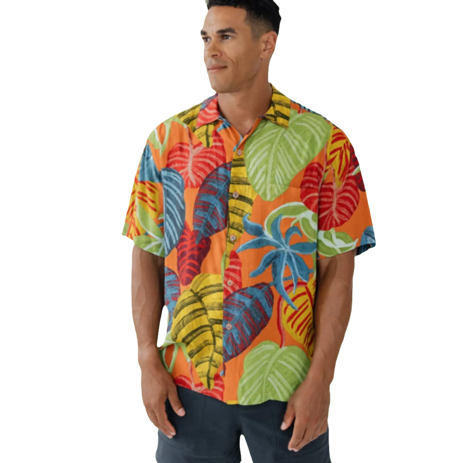 Man stands looking to his right, wearing a short-sleeve button-up shirt set against a white background. The shirt features an extremely vibrant tropical leaf print with a base color of bright orange. The leaves themselves are large and stylized, painted in clashing colors like lime green, bright yellow, royal blue, and deep red. He is wearing dark navy or black shorts with the shirt.