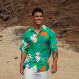 Smiling man stands on a sandy beach with large, tan-colored rock formations rising behind him. He is wearing a short-sleeve button-up shirt featuring a bold, abstract floral print in bright green, white, and orange-red. He pairs the shirt with white shorts or pants. A black tattoo is visible on his left arm, adding to his relaxed, confident look.