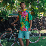 A man stands outdoors next to a dark green bicycle under the shade of a tree. He's wearing a short-sleeve button-up shirt with an extremely colorful, abstract floral pattern that features magenta, red, and green tones. He pairs the shirt with light mint green shorts while resting his hand in his pocket.
