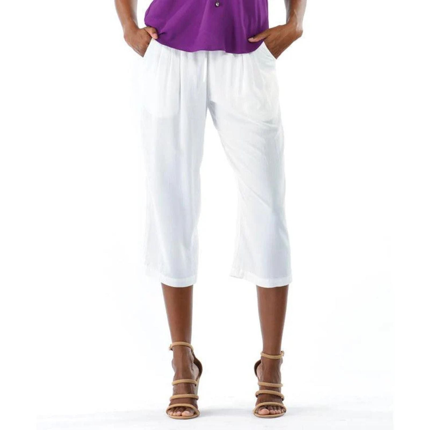 This is a close-up photo of a person wearing the white cropped pants paired with a bright purple top. The person has dark skin and is wearing brown strappy sandals, with their hands tucked into the pants' pockets.