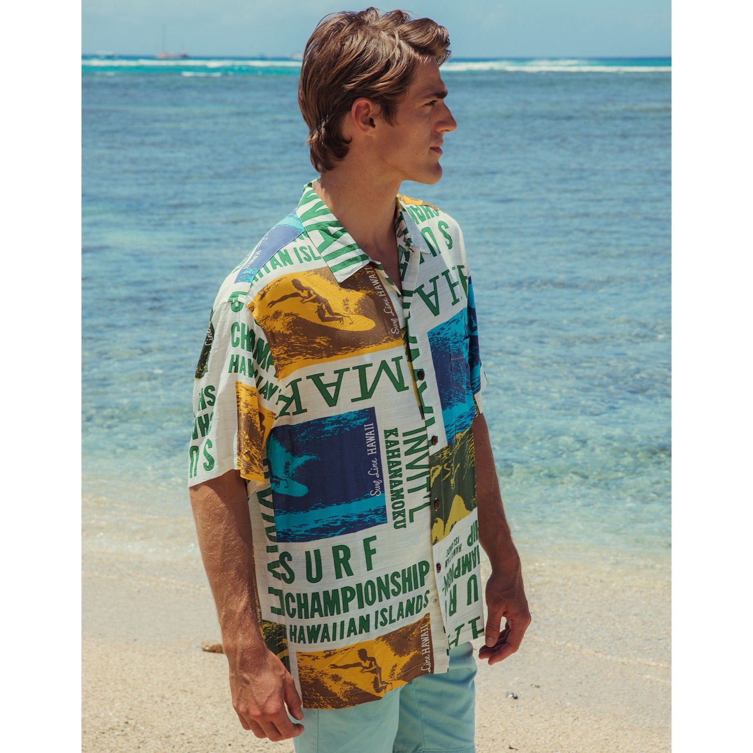 A man wearing the white and multi-colored surf-themed aloha shirt and light blue shorts stands on a sandy beach, looking to the side with the clear, shallow ocean water in the background.