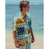 A man wearing the white and multi-colored surf-themed aloha shirt and light blue shorts stands on a sandy beach, looking to the side with the clear, shallow ocean water in the background.