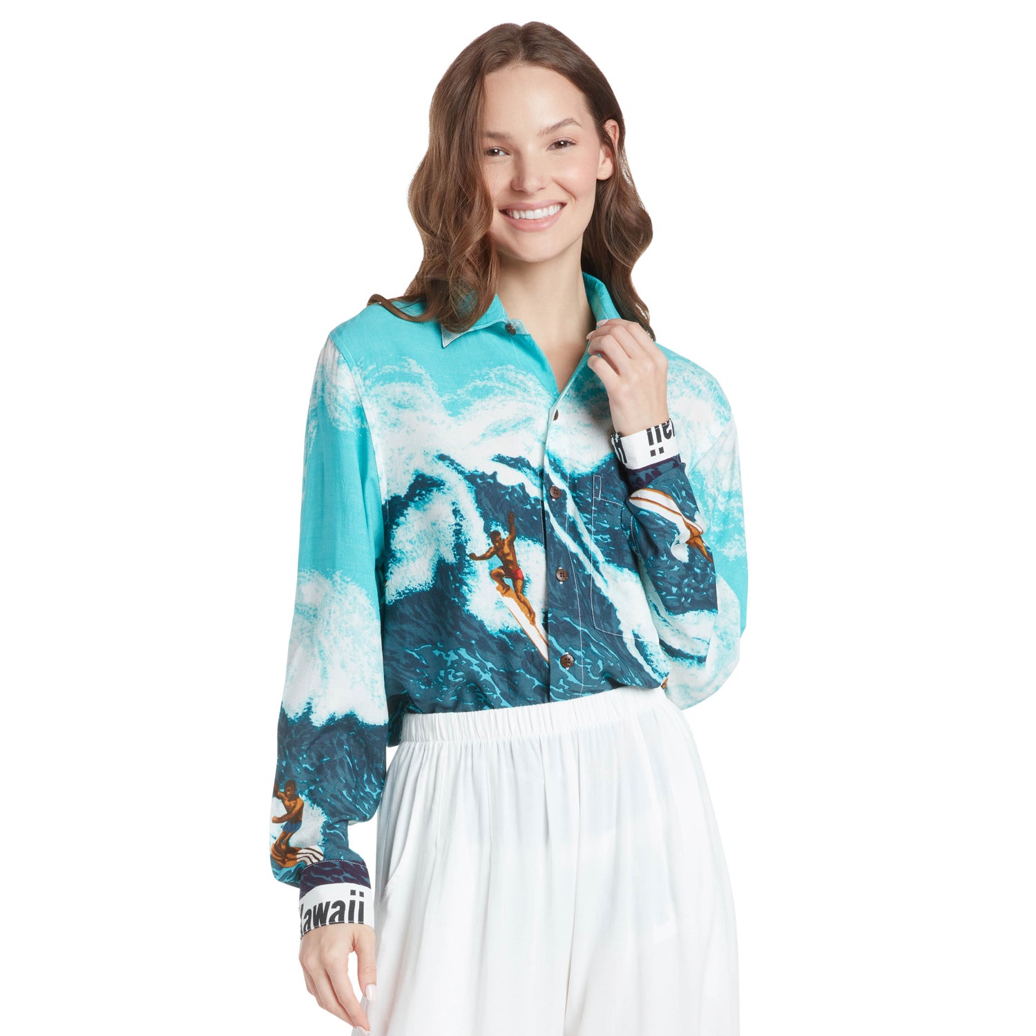 A young woman with brown, wavy hair smiles, touching her collar, while standing against a white background. She is wearing the long-sleeved surfing-print shirt, which depicts blue and white waves and surfers. The "HAWAII" word band is visible on the wide cuffs of her sleeves. She wears the shirt over white wide-leg pants.