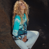 A young woman with long, curly blonde hair sits crouched down on a sandy beach with her eyes closed and face turned up towards the sun, leaning against a dark rock. She is wearing the long-sleeved surfing-print shirt tucked into loose white pants. The word "HAWAII" is visible on the bottom hem and the pants' waistband as she sits.