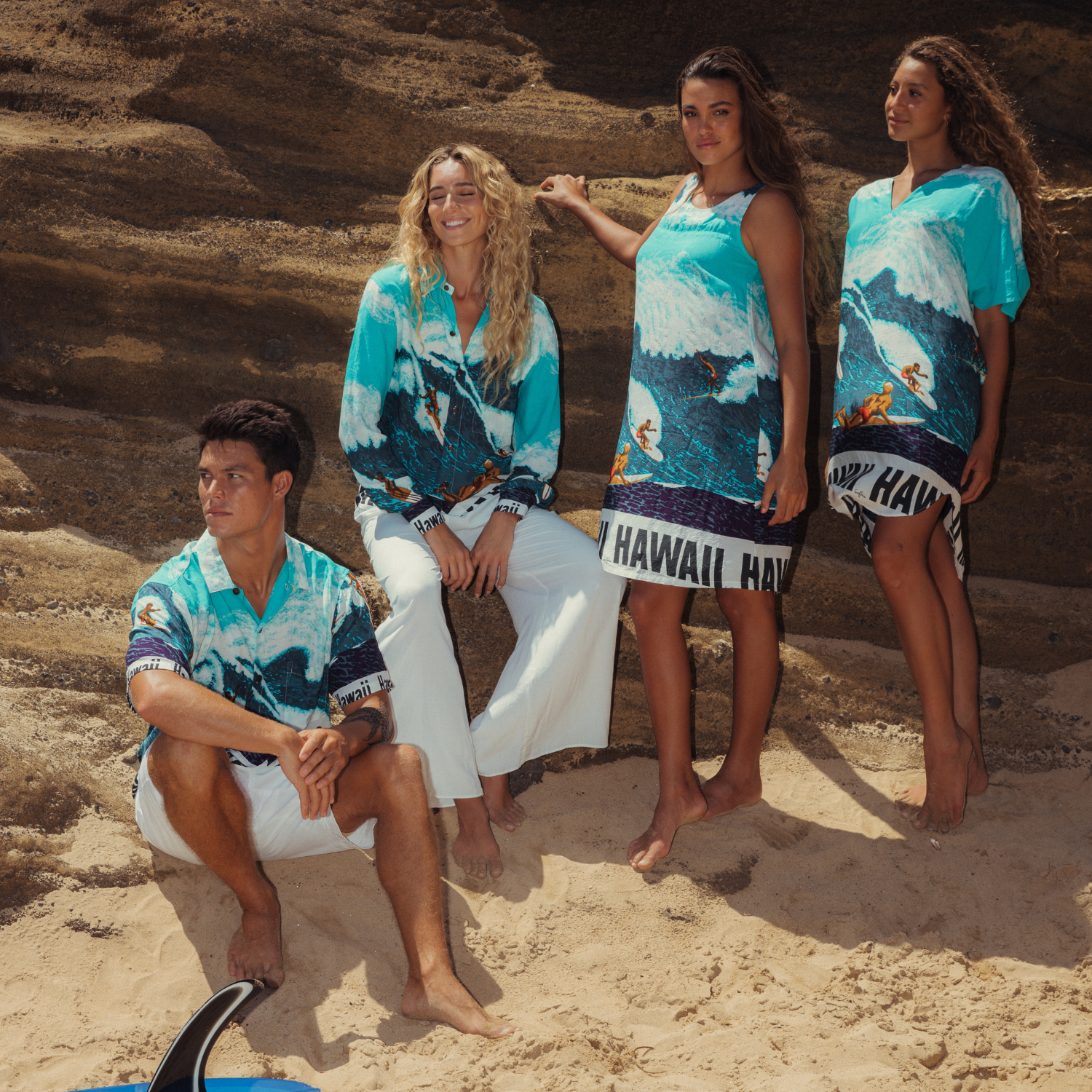 A group of four models posing barefoot on a sandy beach in front of a curved, layered rock formation. They are wearing various garments featuring the same blue, white, and navy surfing-wave print with the "HAWAII" block-letter band. Two women on the right wear dresses, and the woman and man on the left wear shirts and white pants/shorts. A man in the foreground sits near a surfboard fin.
