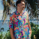 Man stands on a sunny, tropical patch of grass, looking to his right with a slight smile. He is wearing the same vibrant, patchwork-print short-sleeve shirt with dominant fuchsia, blue, and purple tones. The shirt is paired with light khaki shorts. Large palm fronds hand from above, and the blue ocean and bright sky from the backdrop.
