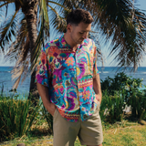Man stands on a grassy patch near the ocean, looking down with a thoughtful expression. He is wearing a short-sleeve button-up shirt featuring an intensely vibrant, multicolored patchwork-style pattern of flowers, birds, and geometric borders. The dominant colors are fuchsia, bright blue, and purple, accented with yellow and orange. He pairs the shirt with solid khaki shorts. Palm trees frame the shot, and the ocean is visible in the background.
