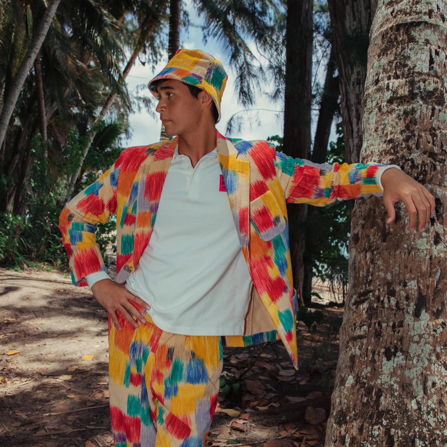 A young man stands in a tropical setting, leaning his left hand on a thick tree trunk, with palm trees visible in the background. He is wearing a colorful, abstract-patterned suit jacket and matching trousers and bucket hat over a plain white polo shirt. The pattern consists of vivid brushstrokes in red, yellow, blue, and green. He looks off to the side, away from the camera.