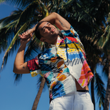 Man stands outdoors on a sunny day with his arms raised and clasped behind his head in a stretching or relaxing pose. He is wearing the same vibrant, abstract-print short-sleeve shirt and white pants. The background is dominated by a clear blue sky and the tall, segmented trunks and fronds of palm trees. A small black tattoo is visible on his upper arm.