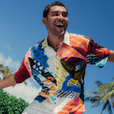 Man stands outdoors under a bright blue sky with his arms slightly outstretched, looking excited or mid-motion. He is wearing a short-sleeve button-up shirt featuring an extremely vibrant, abstract pattern of bold, heavy brushstrokes in colors like fuchsia, royal blue, orange, yellow, and black on a white background. He pairs the shirt with white pants or shorts. The tops of palm trees are visible in the background, suggesting a sunny tropical location.