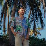 Man stands outdoors under a cluster of palm trees, looking up and to the side. He is wearing the split-print shirt that features both the gray fish and the woven net patterns in tones of blue, green, and brown. He wears the shirt with solid khaki shorts, suggesting a casual, warm-weather style.