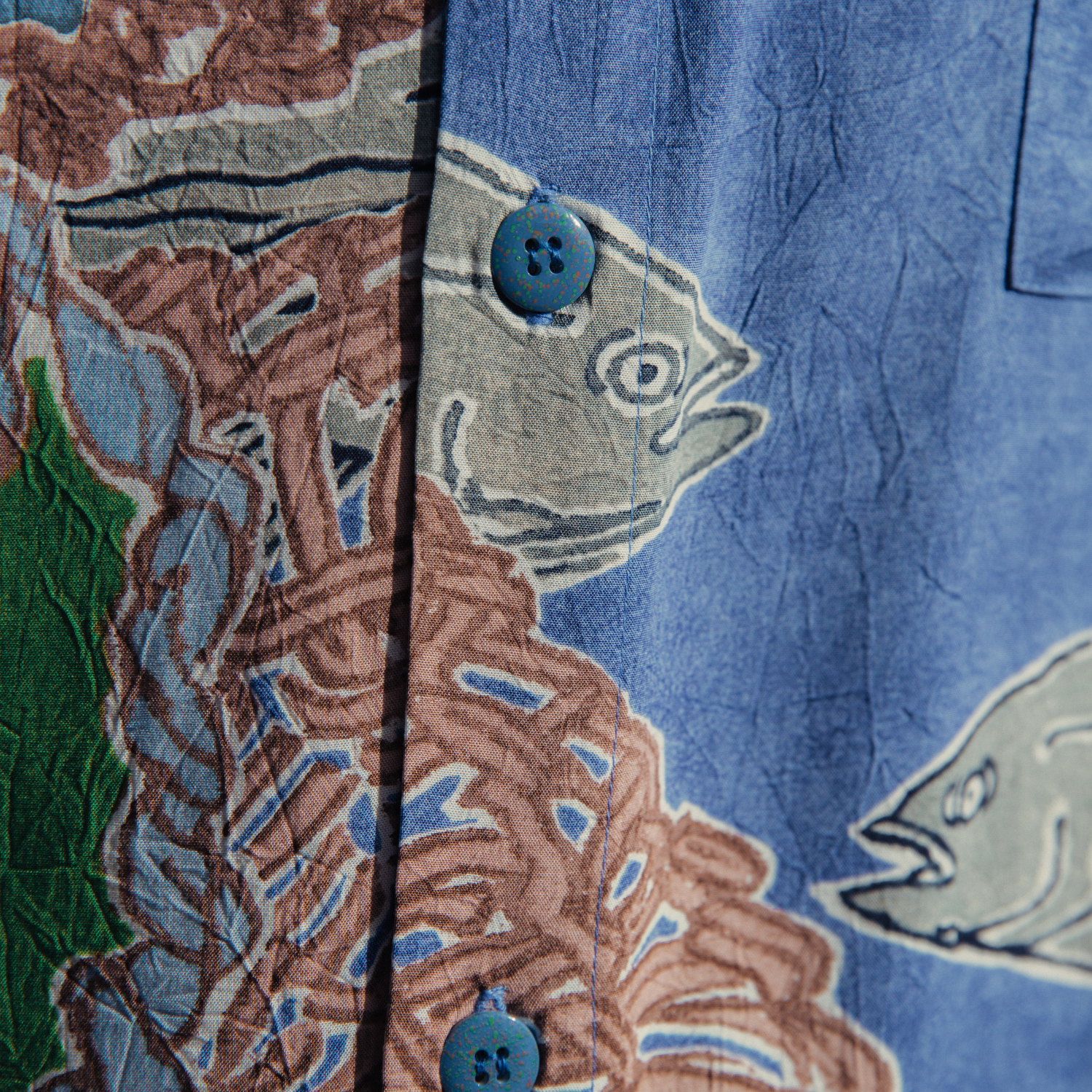 Close-up shot of the shirt's front placket and fabric. The image clearly shows the crinkled texture of the material and the transition between the two patters: the brown woven net design on the left and the gray fish swimming on the blue background on the right. Two small, round blue buttons with a subtle speckled or textured finish are visible on the placket.