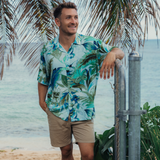 Smiling man stands on a sandy beach in a tropical location, leaning his elbow on a metal gate under the shade of palm fronds. He is wearing a short-sleeved button-up shirt with a vibrant, watery tropical print in shades of deep blue, aqua, and muted olive green on a light blue background. He pairs the shirt with solid khaki shorts. The background shows the ocean and sky meeting on the horizon.