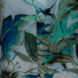 Close-up shot of the shirt's front placket and fabric. The material appears crinkled or textured, clearly showing the blue and green tropical print. The image highlights three small, round buttons on the placket. The buttons are light cream or pale blue and have visible, matching stitching and a slightly textured surface.