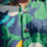Close-up shot of a garment, showing the upper chest and collar area. The fabric is a crinkled, textured material in a bold abstract floral pattern featuring dark navy blue, lime green, bright turquoise, white, and yellow shapes. The garment is buttoned up, showing two dark brown, two-hole buttons on the front placket, with a hint of a thin gold chain visible around the person's neck under the collar.