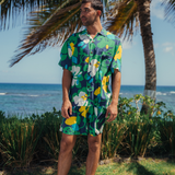 Man stands outdoors wearing a matching short-sleeve button-up shirt and shorts set in the same abstract, dark-toned floral print (navy blue, green, white, yellow, and turquoise). He is positioned on a grassy lawn in a tropical location, looking off to the side. Behind him, there's a large palm tree, and in the background, a bright blue ocean meets the horizon under a sunny sky.