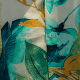 Close-up shot of the shirt's front placket and fabric texture. The material appears crinkled or textured and clearly displays the watercolor-style tropical pattern with large swaths of teal, dark green, and yellow over a light background. The image clearly shows a small, round button on the placket, which is the same teal-green color as the fabric and has a subtly textured, etched surface.