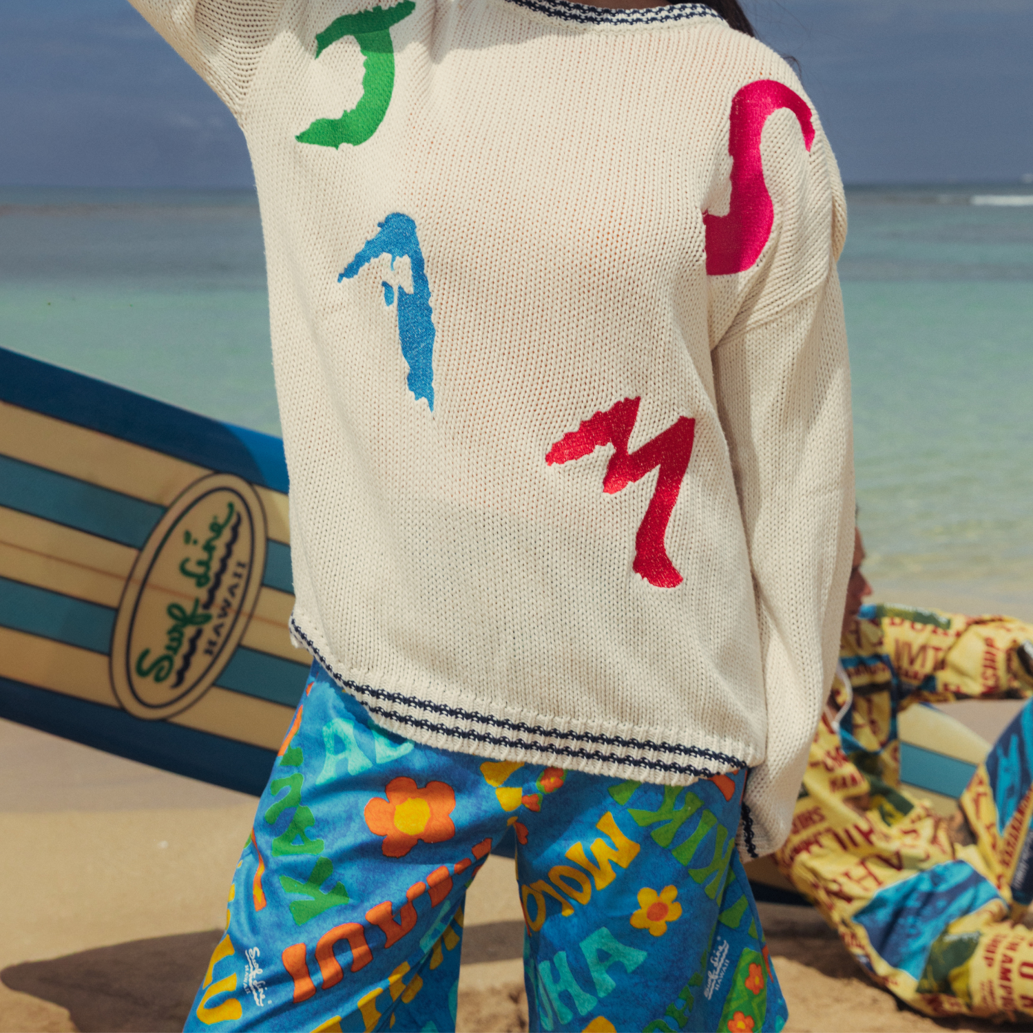 Person stands on a sandy beach with their back mostly to the camera, wearing a cream-colored knit sweater over brightly patterned blue shorts. The sweater features large, abstract, colorful patches that spell out "JAMS" on the back in green, blue, pink, and red. The person is standing next to a striped surfboard partially visible on the sand, and the background shows a light blue ocean meeting a bright sky. Another person is partially visible, lying down in the sand wearing colorful patterned clothing.