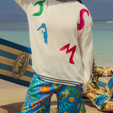Person stands on a sandy beach with their back mostly to the camera, wearing a cream-colored knit sweater over brightly patterned blue shorts. The sweater features large, abstract, colorful patches that spell out "JAMS" on the back in green, blue, pink, and red. The person is standing next to a striped surfboard partially visible on the sand, and the background shows a light blue ocean meeting a bright sky. Another person is partially visible, lying down in the sand wearing colorful patterned clothing.