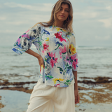 Woman wearing a colorful floral blouse and white pants standing on a beach.