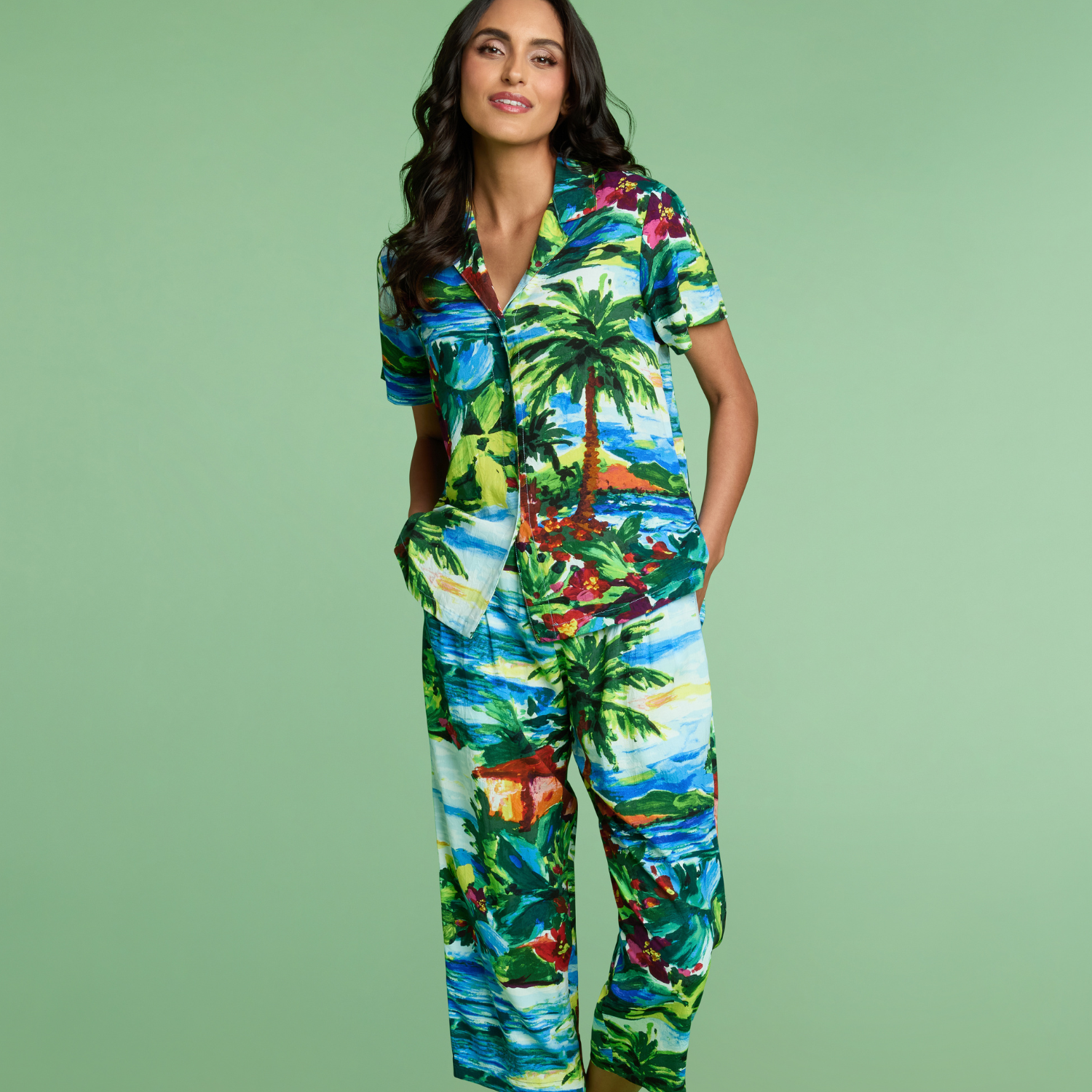 Woman wearing a tropical-themed button down shirt with color and pants of the same print on a green background