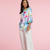 Woman wearing a colorful floral top and white pants on a pink background