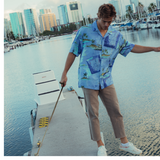This is a photo of a young man wearing the blue nautical patterned shirt while standing on a concrete pier next to a marina. He is looking down and is wearing cropped tan pants and white sneakers. The background shows a city skyline across the water, with numerous sailboats and yachts docked nearby.