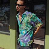 A young man wearing dark sunglasses and the short-sleeved floral shirt stands in profile on a sunny street in front of a lime-green storefront. The shirt's vibrant blue, green, and purple floral pattern contrasts with the bright green building. He is wearing dark gray or black jeans and has his hands on his hips, looking off to the side.
