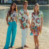 This is a lifestyle photo showing three young models standing barefoot on a sandy beach in front of a body of water. They are all wearing clothing with the same orange and blue floral pattern: a woman on the left in a tied-front shirt and turquoise pants, a man in the center in a button-up shirt and light pants, and a woman on the right in a short dress with a colorful fringed hem.