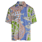Product shot of a short-sleeve button-up shirt laid flat against a white background. The shirt features a two-panel design with a split print. The right side and sleeve show gray fish swimming against a solid blue background. The left side and sleeve feature an abstract pattern of brown and green woven nets or traps, also against a blue background. The overall color scheme is blue, green, brown, and gray.