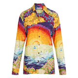 A long-sleeved button-down shirt displayed on white background. The fabric features a vibrant, painterly scene of a sunset over the ocean. The top half shows a large, radiating orange and yellow sun setting against a sky of red, purple, and blue, framed by bright yellow and blue flowers. The bottom half depicts a sparkling blue and yellow ocean with small white sailboats, and a shoreline lined with pink and green flowers. The shirt has brown buttons down the front.