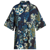 A short-sleeved, dark blue batik-style Hawaiian shirt with an abstract pattern of cream, light blue, and moss green floral and geometric shapes. It has a camp collar and brown buttons.