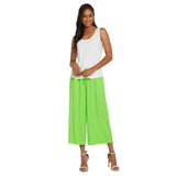 A smiling woman with long dark hair is standing on a white background, wearing a simple white sleeveless tank top tucked into the same bright lime green wide-leg cropped pants, and completing the look with tan high-heeled sandals.