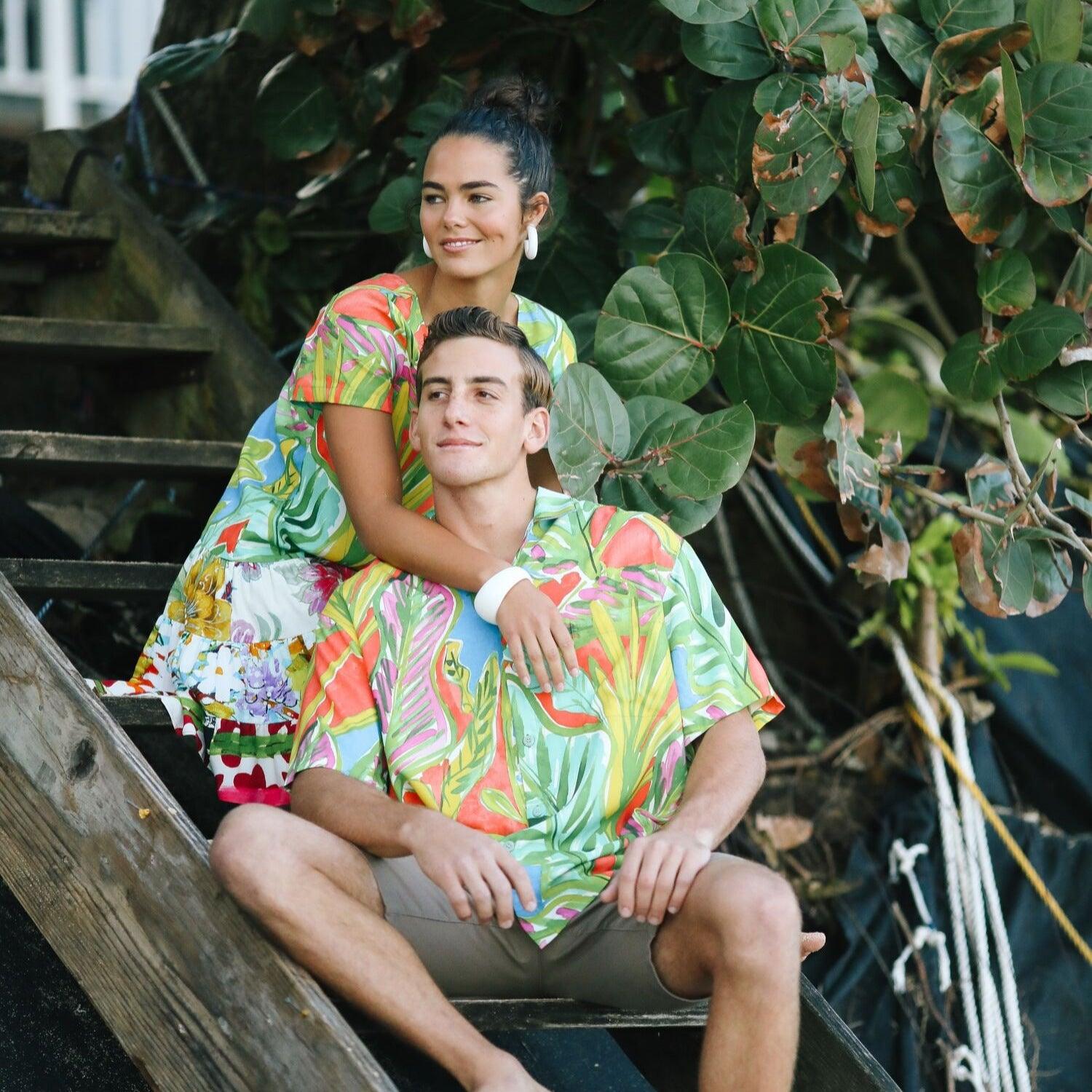 This outdoor photo shows a couple sitting on wooden stairs under lush green foliage, both wearing garments with similar vivid tropical prints. The man is wearing the short-sleeved shirt with khaki shorts, while the woman behind him wears a dress, with her arm resting on his shoulder.