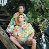 This outdoor photo shows a couple sitting on wooden stairs under lush green foliage, both wearing garments with similar vivid tropical prints. The man is wearing the short-sleeved shirt with khaki shorts, while the woman behind him wears a dress, with her arm resting on his shoulder.