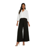 A woman with long dark hair is smiling and standing on a white background, wearing a white, three-quarter sleeve collared shirt tucked into the same black wide-leg trousers and accessorized with tan high-heeled sandals.