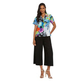 A woman with long dark hair is smiling and standing on a white background, wearing a short-sleeve collared shirt with a vibrant, multicolored tropical floral print over loose-fitting, black wide-leg cropped pants and tan high-heeled sandals.