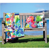 A wooden slatted bench sits on a bright green lawn overlooking the ocean, draped with a colorful patchwork blanket and decorated with two throw pillows: one heart-shaped and one square, all featuring coordinating floral and abstract prints.