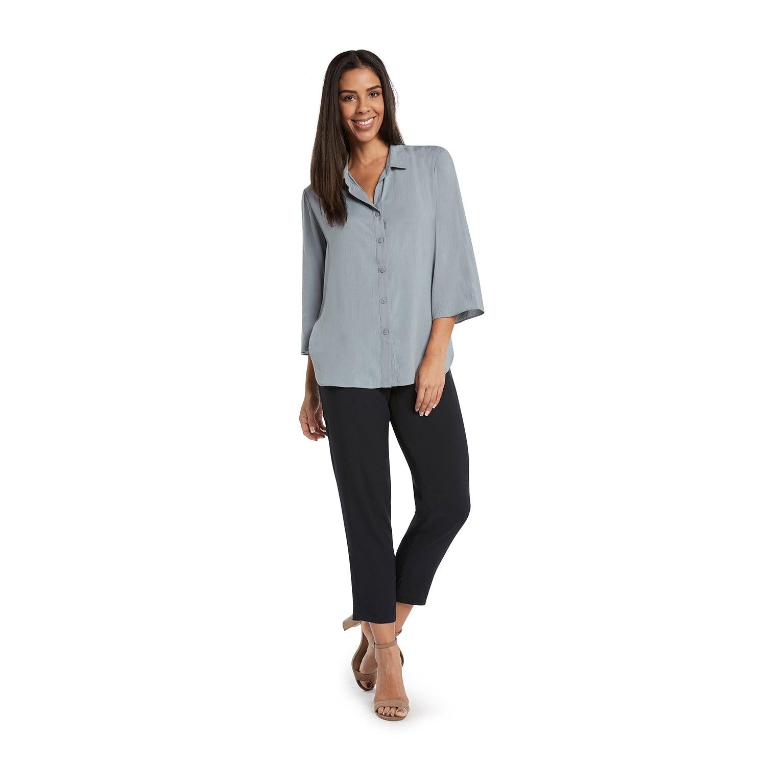 A woman with long dark hair smiles while standing on a white background, wearing a light blue-gray, three-quarter sleeve collared blouse and fitted black cropped pants with tan open-toed heels.