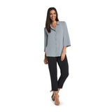 A woman with long dark hair smiles while standing on a white background, wearing a light blue-gray, three-quarter sleeve collared blouse and fitted black cropped pants with tan open-toed heels.