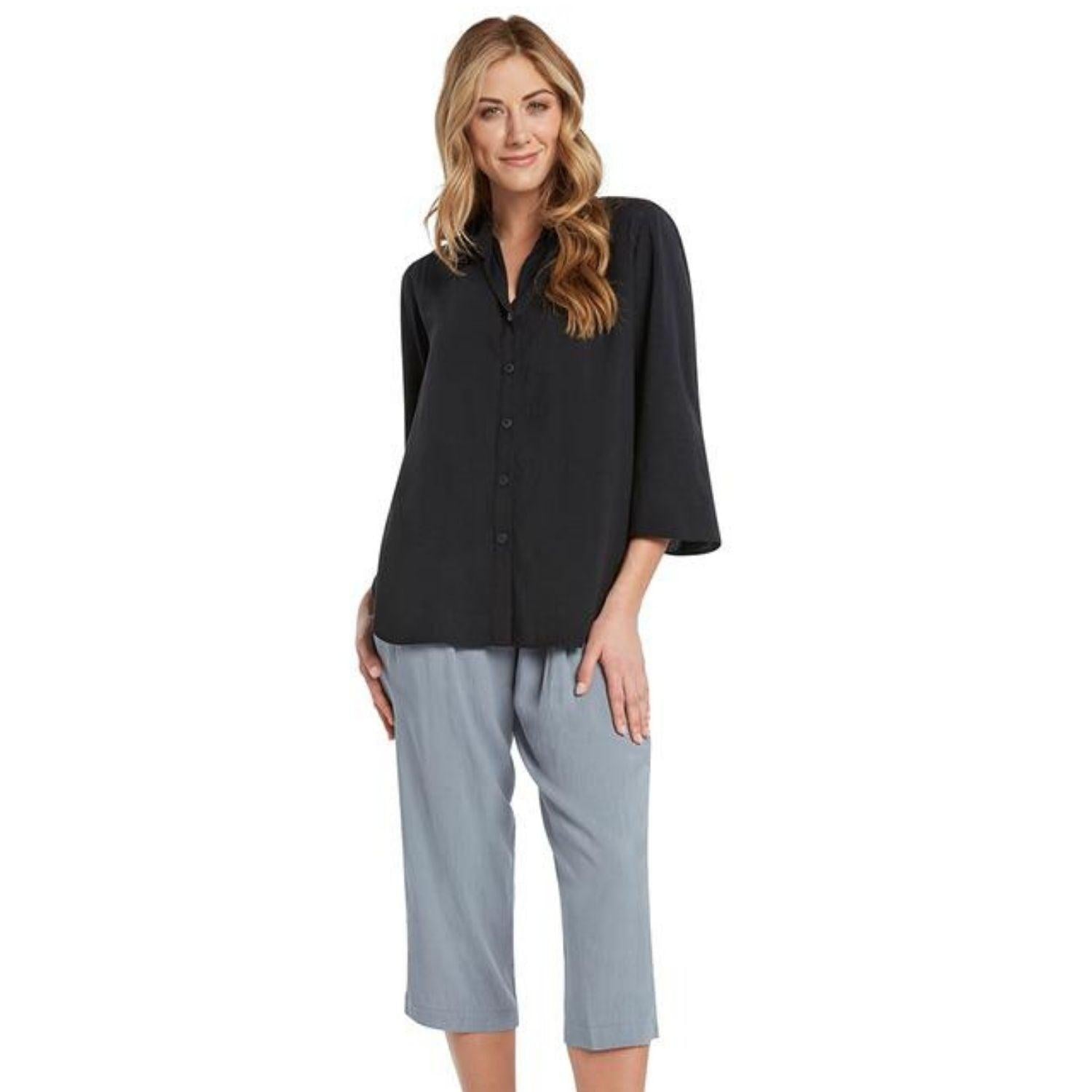 A blonde woman with wavy hair stands on a white background, wearing a loose, black collared blouse with three-quarter bell sleeves tucked into relaxed-fit, light blue-gray cropped pants.