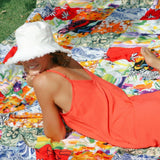 A woman wearing a bright orange sundress and a white frayed-brim bucket hat is lying on her stomach on a vividly colorful patchwork blanket in a grassy area, resting her head on her hand and smiling.