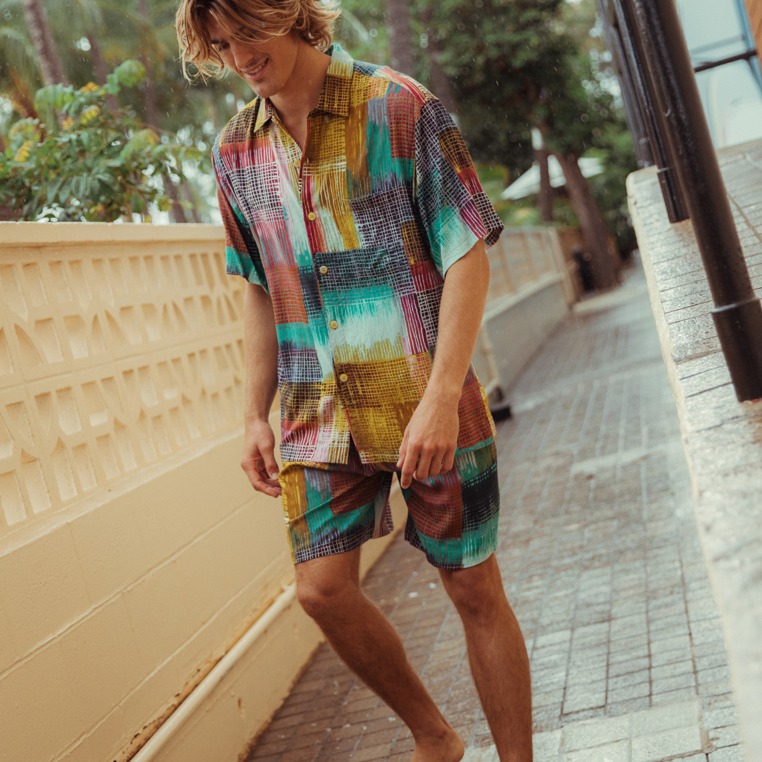 A young man with long blonde hair is walking down a narrow outdoor walkway flanked by yellow walls, while smiling and wearing a matching short-sleeve shirt and shorts set that features the same vibrant, abstract, multicolored patchwork print.