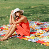 A woman with a short hairstyle and a white brimmed hat sits barefoot on a large, colorful patchwork blanket laid out on a green grassy lawn; she is wearing a bright coral-red sundress and is leaning forward with her elbows resting on her knees.