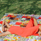 A woman in a coral-red jumpsuit and a white hat is lying on her back, stretching her arms above her head on a brightly colored patchwork blanket on a sunny patch of grass, with her eyes shielded from the sun.