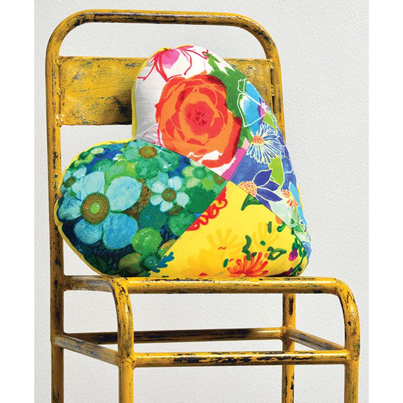 This is a heart-shaped pillow resting on a distressed yellow metal chair. The pillow is made from a patchwork of colorful floral and abstract prints, including a large orange rose, bright blue flowers, and green leaves.