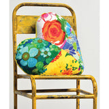 This is a heart-shaped pillow resting on a distressed yellow metal chair. The pillow is made from a patchwork of colorful floral and abstract prints, including a large orange rose, bright blue flowers, and green leaves.