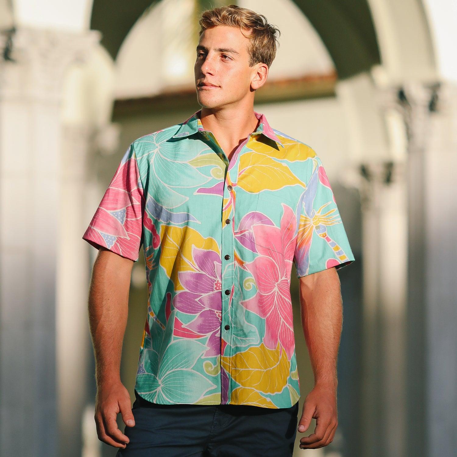 This outdoor photo shows a young man wearing a short-sleeved shirt with a vibrant floral and leaf pattern in shades of jade green, pink, yellow, and purple. He is standing in front of light-colored architectural arches, looking off to his right with his hands down at his sides.
