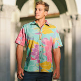 This outdoor photo shows a young man wearing a short-sleeved shirt with a vibrant floral and leaf pattern in shades of jade green, pink, yellow, and purple. He is standing in front of light-colored architectural arches, looking off to his right with his hands down at his sides.